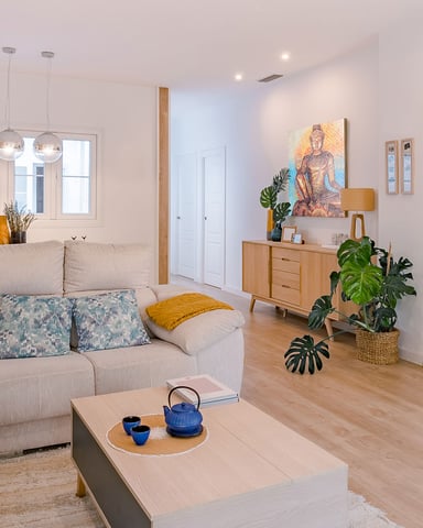 Bright open-plan living room with beige sectional sofa, wooden coffee table, flat-screen TV, and multiple potted plants throughout