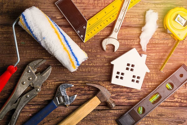 Home improvement tools and supplies arranged on wooden surface: paint roller, tape measure, wrench, hammer, level, and white house-shaped object.