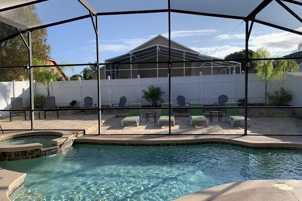 Screened pool area with clear turquoise water, lounge chairs, and spa, overlooking white fence and covered lanai structure