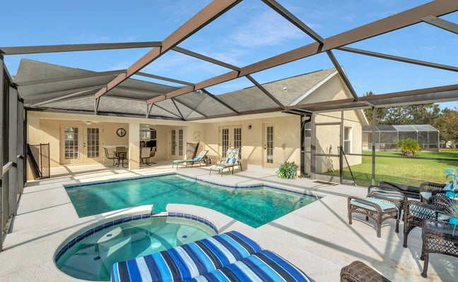 Screened pool and spa area with tan house, metal pergola, lounge chairs, and fenced yard beyond
